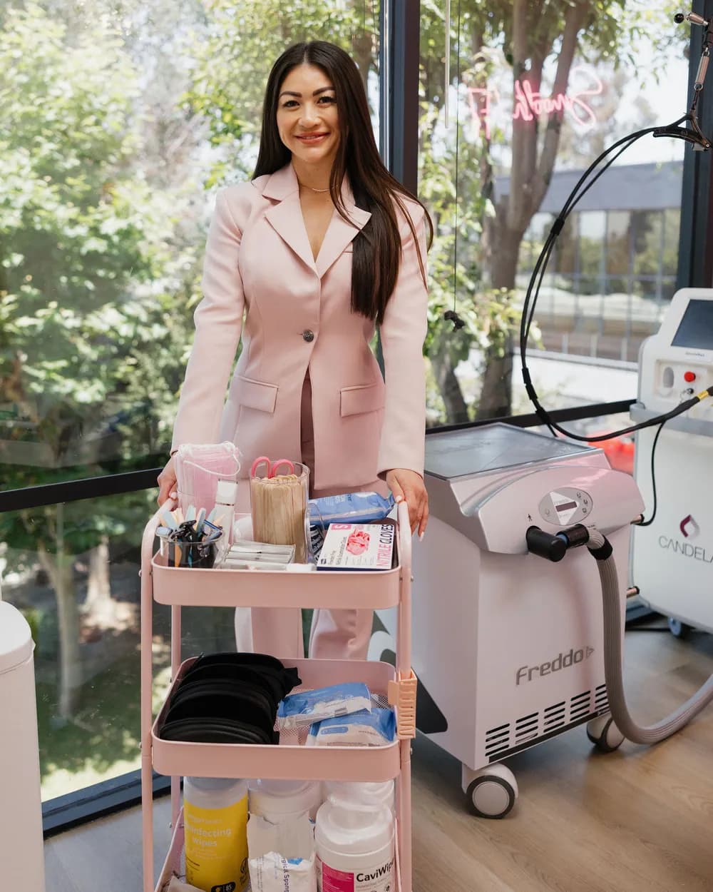 Leslie in a blush suit behind the pink supply cart in her treatment room, with the Candela laser and Freddo cooler beside her