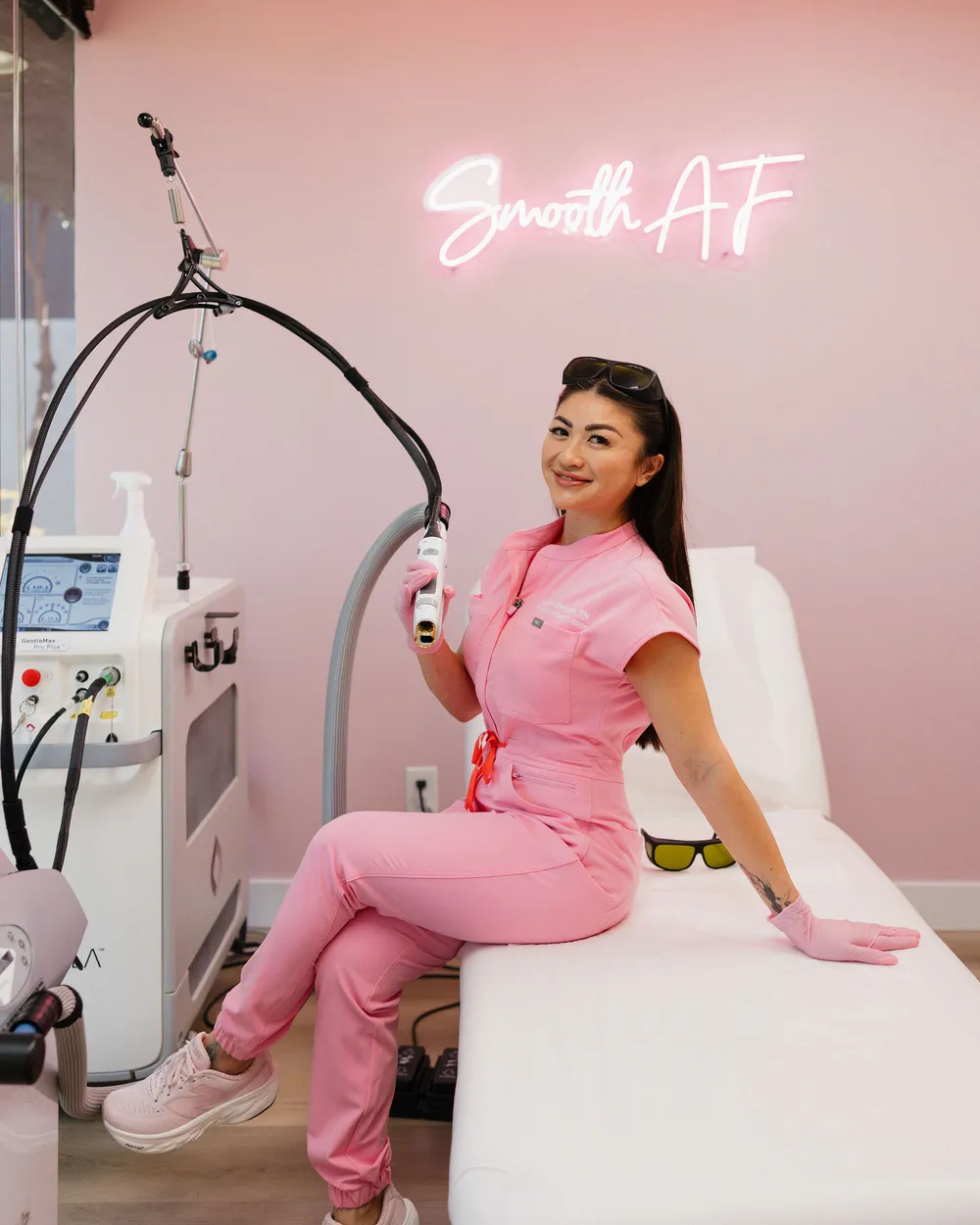 Leslie seated on the treatment bed holding the Candela GentleMax Pro handpiece, the Smooth AF neon behind her