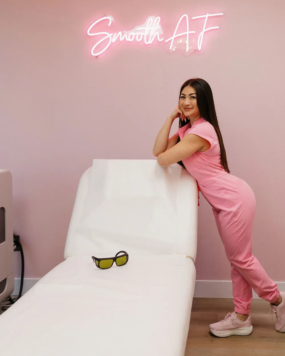 The Pink Diamond treatment room: Leslie leaning on the treatment bed under the Smooth AF neon, laser goggles resting on the paper